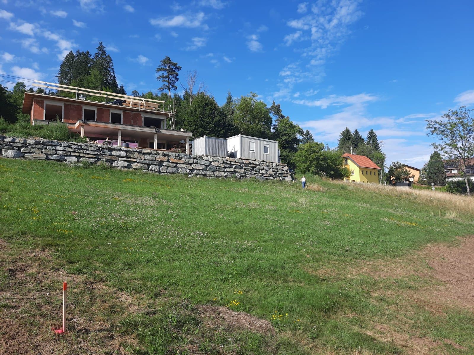Das Grundstück in Köttmannsdorf: eine Wiese am Hang mit einem Vermessungspflock im Vordergrund, dahinter eine Steinmauer und Häuser.