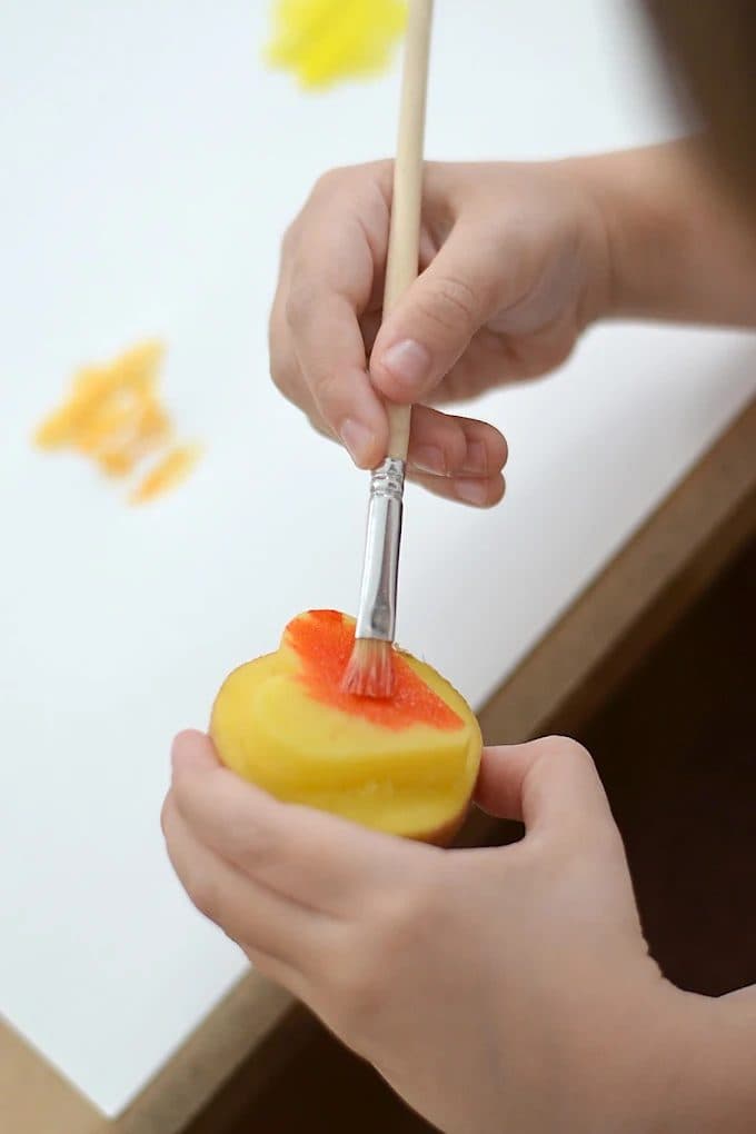 Kinderhände bemalen einen Kartoffelstempel mit oranger Farbe