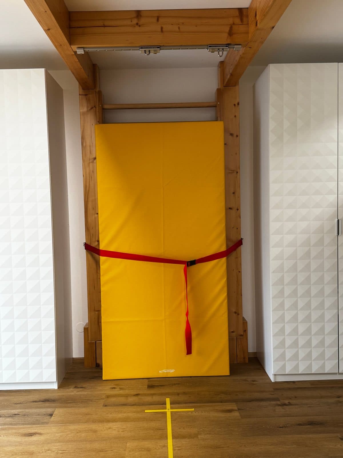Sprossenwand aus Holz mit gelber Turnmatte zwischen weißen Schränken