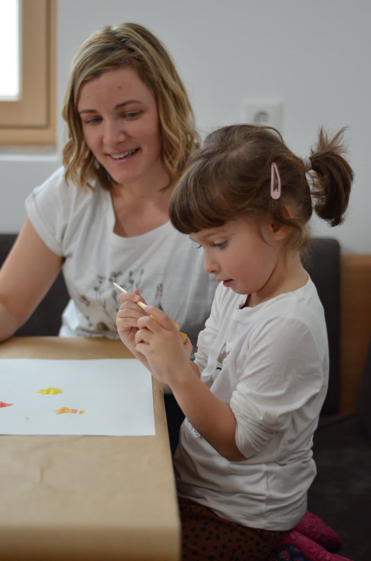 Lisa Lugger Breidler sitzt neben einem Mädchen, das mit einem Holzstäbchen Farbe auf ein Blatt Papier tupft